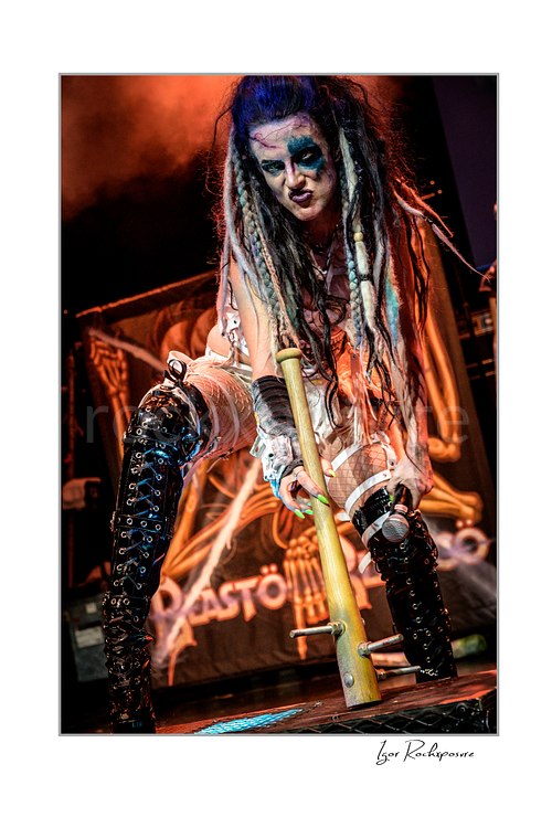 Vertical color image of Calico Cooper of Beasto Blanco in a fierce live performance pose, holding a microphone and gripping a stage prop with dramatic makeup, long dreadlocks, and warm stage smoke behind her