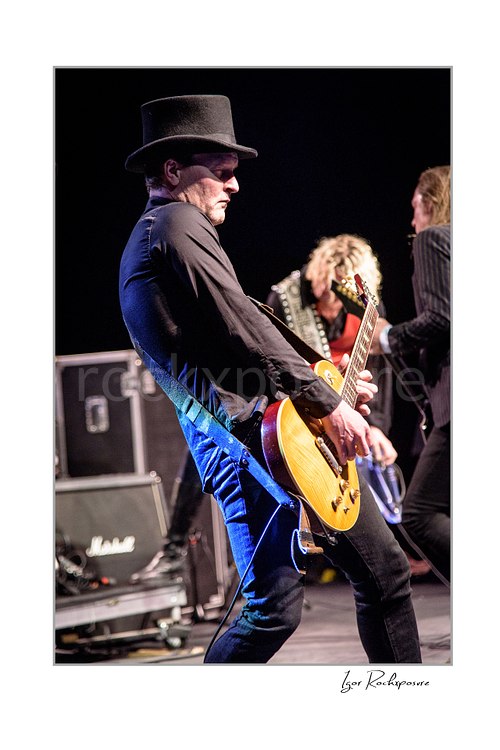 Vertical color image of Jacob Binzer playing guitar on stage in a dark hat, with dramatic stage light and blurred band members in the background