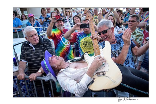 Horizontal color image of Randy Hansen playing a white guitar while leaning back into the crowd during an outdoor ship deck performance