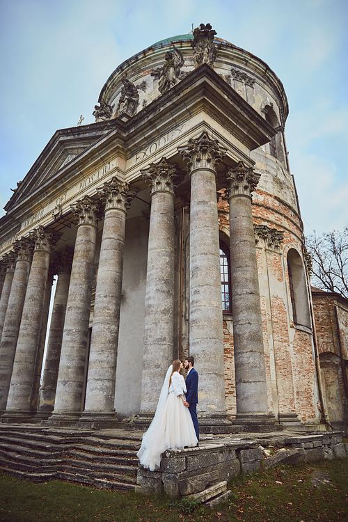 Bride and groom beneath the historic church columns in Pidhirtsi