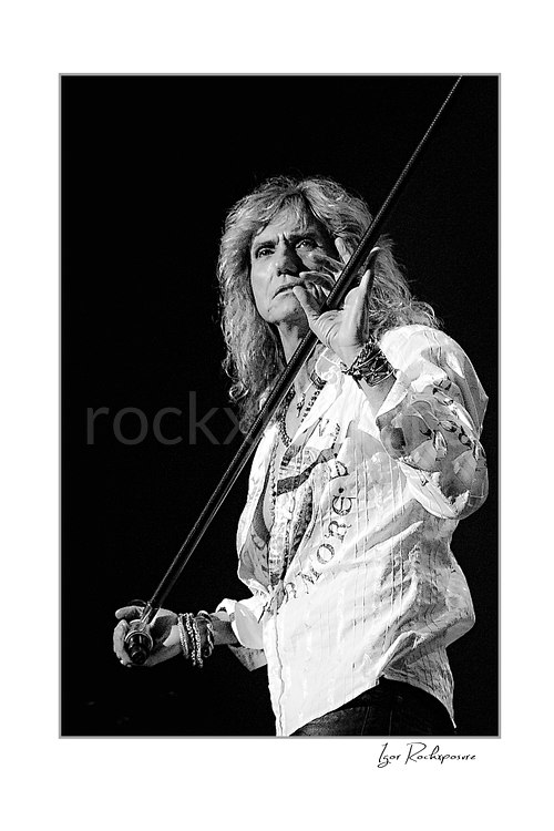 Vertical black and white image of David Coverdale of Whitesnake performing live on stage, holding a microphone stand against a dark background