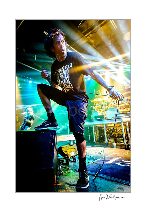 Vertical color image of Randy Blythe of Lamb of God performing on stage with one foot raised, handheld microphone, and bright concert beams