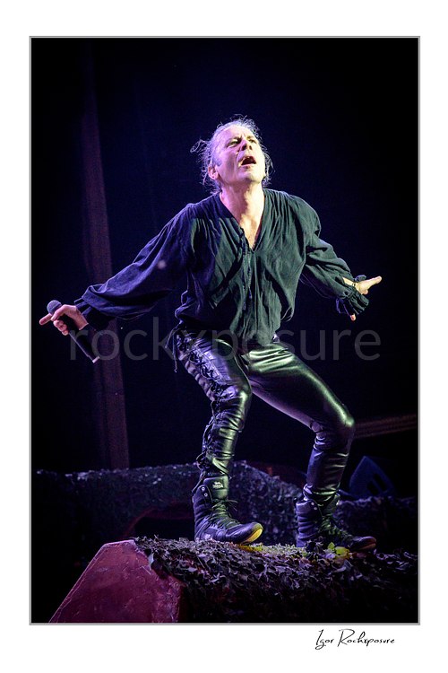 Vertical color image of Bruce Dickinson of Iron Maiden performing live on stage, holding a microphone and striking an expressive pose under concert lighting.