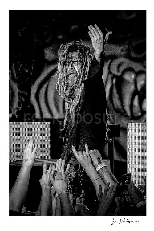 Vertical black and white image of Rob Zombie performing live on stage above the crowd with long dreadlocks, raised hand, and fans reaching toward him
