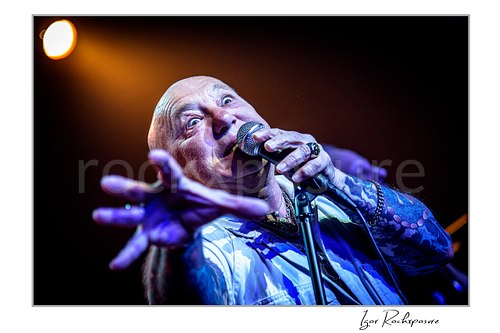 Horizontal color image of Angry Anderson of Rose Tattoo singing into a microphone in a dramatic close-up with an outstretched hand under blue and warm concert light