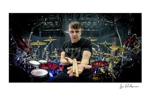 Horizontal color image of Korn drummer Ray Luzier seated at his drum setup before the show, looking at the camera with drumsticks balanced across his open hand
