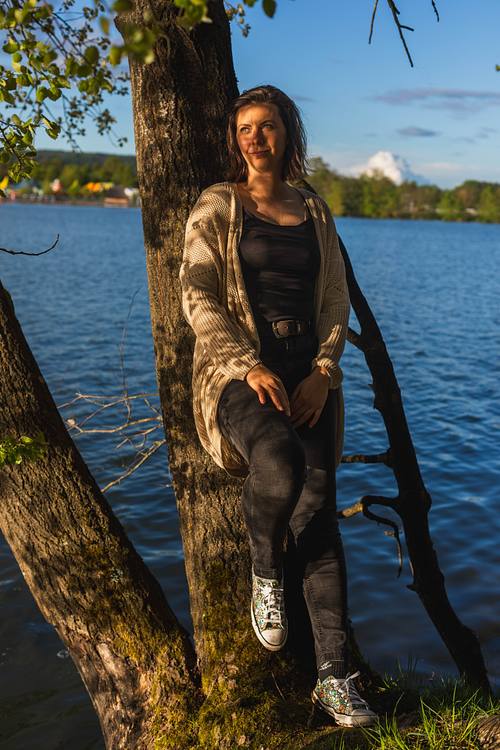 Eine Frau in Jeans, Sneakern und langem Cardigan lehnt entspannt an einem Baumstamm direkt an einem blauen See im Abendlicht.