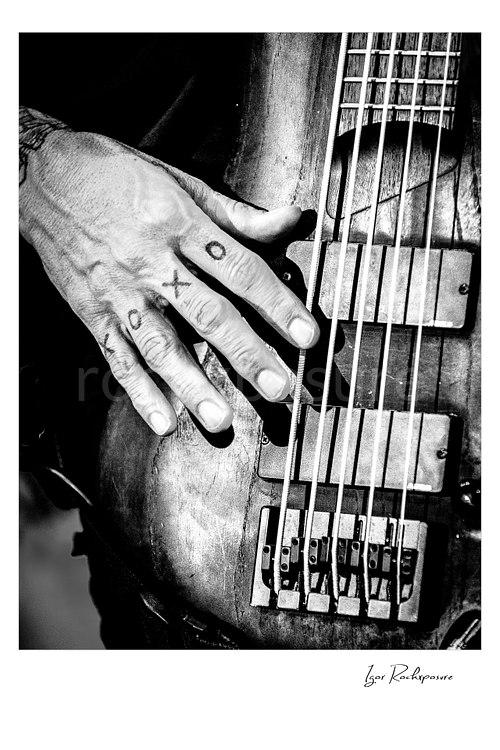 Vertical black and white image of Korn bassist Fieldy’s tattooed hand resting on the strings of a bass guitar in a tight close-up