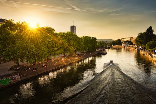 Fotografie des Wiener Donaukanals im warmen Gegenlicht der untergehenden Sonne. Auf dem Fluss fährt ein Boot und zieht eine markante, V-förmige Heckwelle durch das glitzernde Wasser. Am linken, von dichten grünen Bäumen gesäumten Ufer sitzen zahlreiche Menschen auf den Stufen und genießen die frühsommerliche Abendstimmung. Im Hintergrund ist die Silhouette der Wiener Stadtlandschaft sichtbar.
