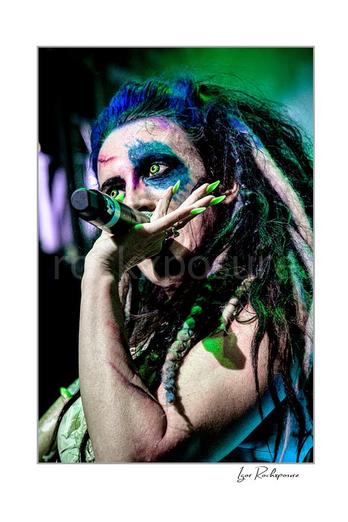 Vertical color image of Calico Cooper of Beasto Blanco singing into a microphone in a tight live performance close-up, with dramatic eye makeup, dreadlocks, and neon green nails under green stage light