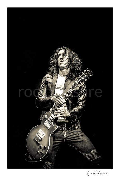 Vertical black and white image of Brent Woods playing guitar on stage in a dramatic full-body performance shot