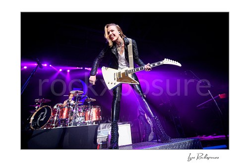 Horizontal color image of Lzzy Hale of Halestorm performing live with a white guitar at the front of the stage, smiling under vivid purple concert light with Arejay Hale visible at the drum kit behind her