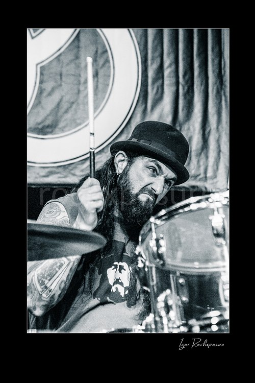 Vertical black and white image of Mike Portnoy at the drum kit wearing a bowler hat and raising a drumstick with an intense expression