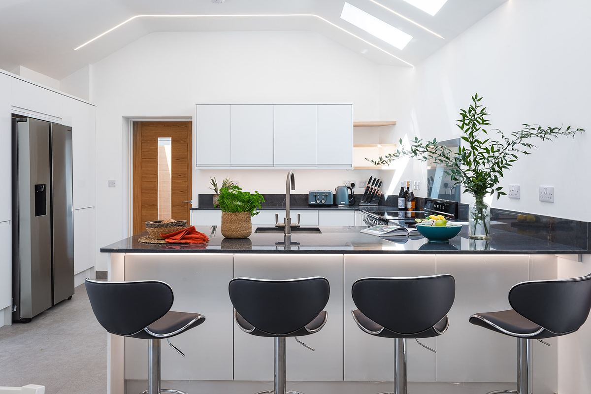 Modern white kitchen with a central island, four grey bar stools, stainless steel appliances, and potted plants on the counter. Perfect for holiday home photography Cornwall, with natural light streaming in from skylights above.