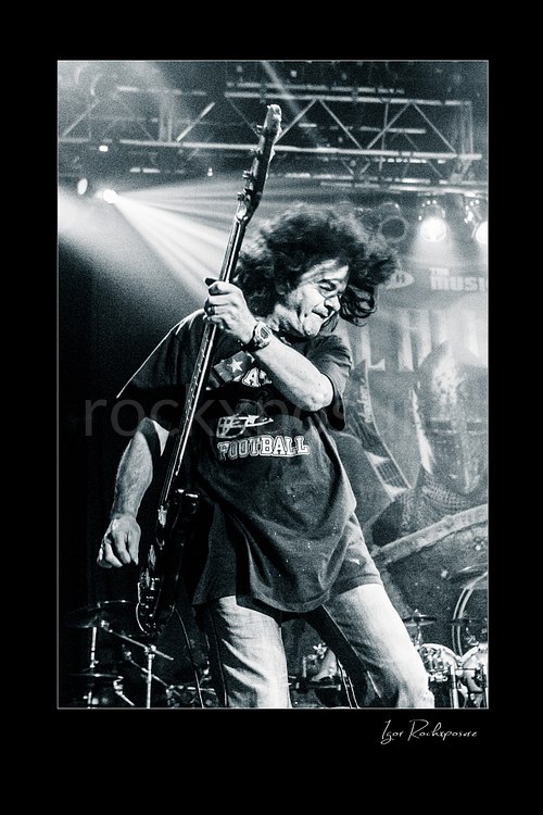 Vertical black and white image of Jimmy Bain playing bass onstage in an energetic live performance shot