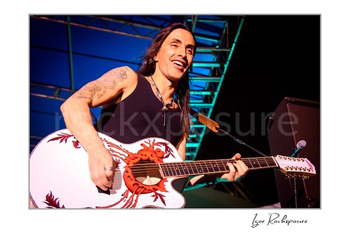 Horizontal color image of Nuno Bettencourt smiling during a live performance with a white acoustic guitar featuring a bold red graphic design