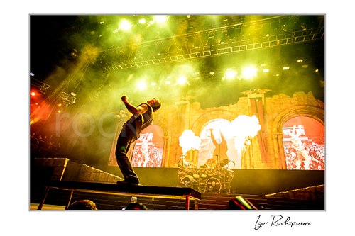 Horizontal color image of M. Shadows of Avenged Sevenfold leaning back on stage with fire bursts, stage smoke, and vivid green concert lights