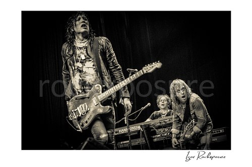Horizontal black and white image of Tom Keifer, Paul Taylor, and Tony Higbee performing live on stage, with Keifer in the foreground while Taylor plays keys and Higbee leans into his guitar beside him