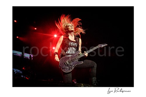 Horizontal color image of Nita Strauss performing live on stage with an electric guitar and dramatic flying hair under red stage lighting