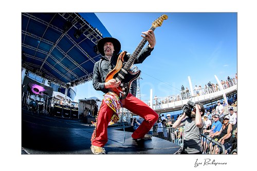 Horizontal color image of Randy Hansen playing guitar in a wide stage stance on an outdoor ship deck stage under bright daylight