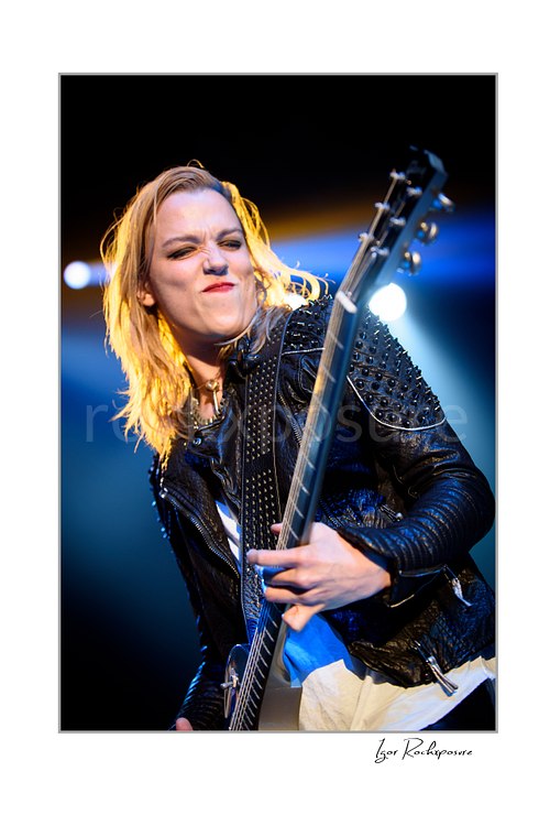 Vertical color image of Lzzy Hale of Halestorm playing guitar in a fierce live performance close-up, with a studded black jacket, intense expression, and bright blue-white stage lights behind her