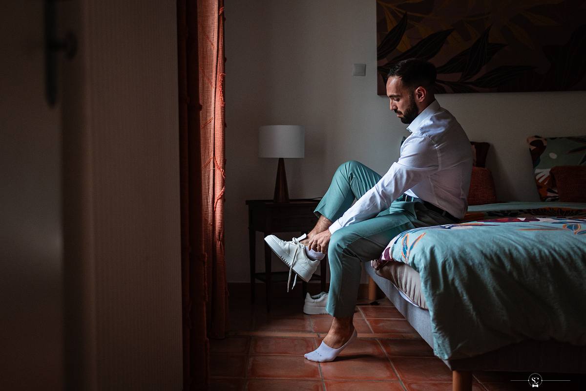 Une pr&eacute;paration soign&eacute;e pour le grand jour, montrant le mari&eacute; assis au bord d'un lit, enfilant avec soin des baskets blanches, dans une chambre &agrave; l'ambiance paisible, &eacute;voquant l'esprit du mariage simple &agrave; N&icirc;mes