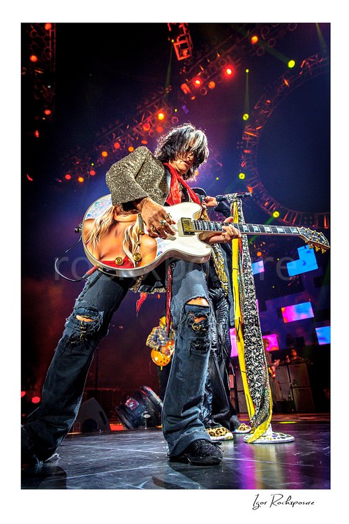 Vertical color image of Joe Perry of Aerosmith performing live on stage with a custom painted guitar, glitter jacket, ripped jeans, and scarf-wrapped microphone stand