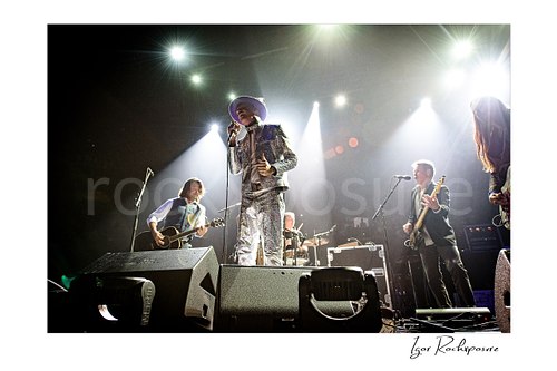 Horizontal color image of The Tragically Hip performing live on stage, with Gord Downie in a glittering silver suit at center under bright concert spotlights and band members visible across the stage