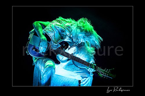 Horizontal color image of John 5 performing live on stage under blue and green lighting with a black electric guitar, white studded stage outfit, and flying hair