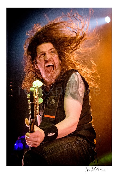 Vertical color close-up image of Frank Bello of Anthrax performing live on stage, playing bass guitar with his hair flying under warm concert lighting
