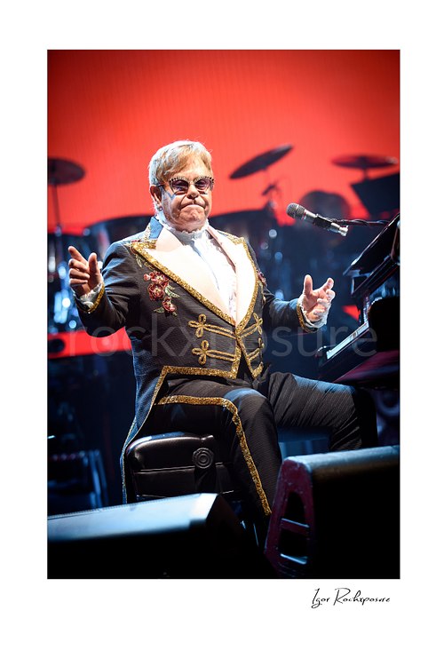 Vertical color image of Elton John seated at the piano with arms outstretched against a vivid red stage backdrop