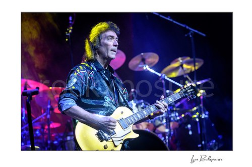 Horizontal color image of Steve Hackett performing live with electric guitar on stage with the drum kit behind him