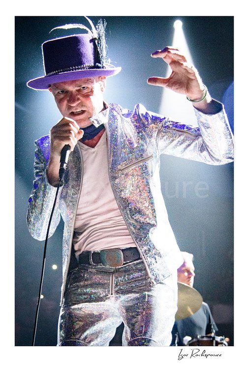 Vertical color image of Gord Downie of The Tragically Hip singing into a microphone in a dramatic close live performance shot, wearing a glittering silver suit and hat with bright stage light behind him