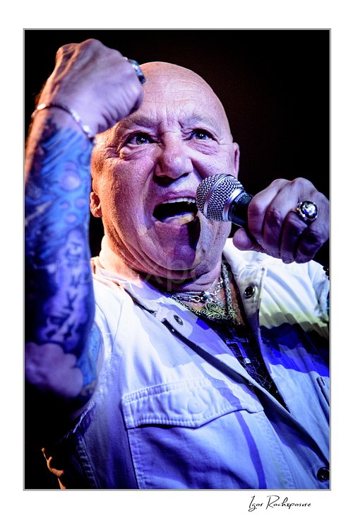 Vertical color image of Angry Anderson of Rose Tattoo singing into a microphone in a tight close-up with raised fist under blue and warm concert light
