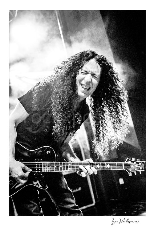 Vertical black and white image of Marty Friedman playing guitar onstage with long curly hair and intense expression under bright stage light