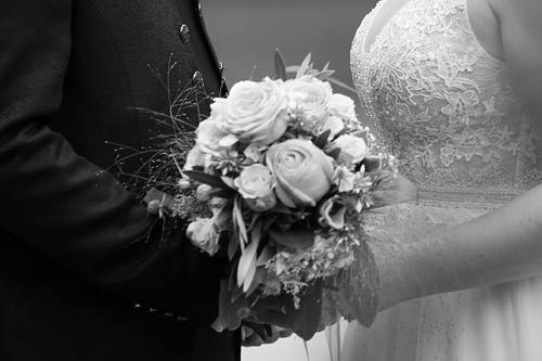 Detailaufnahme in Schwarz-Weiß: Ein Brautstrauß wird von dem Brautpaar gemeinsam gehalten, im Anschnitt sind die Hochzeitskleidung und Hände zu sehen.