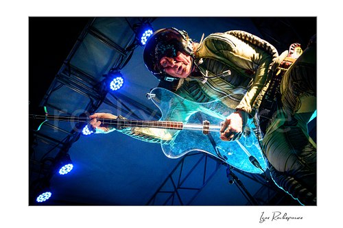 Horizontal color image of Stig Pedersen of D-A-D performing live in a metallic space suit and helmet, photographed from below with his clear bass guitar extending toward the camera under blue stage lights