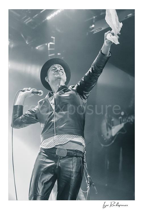 Vertical black and white image of Gord Downie of The Tragically Hip holding a microphone and lifting a white cloth high on stage, wearing a hat and leather jacket in bright concert haze