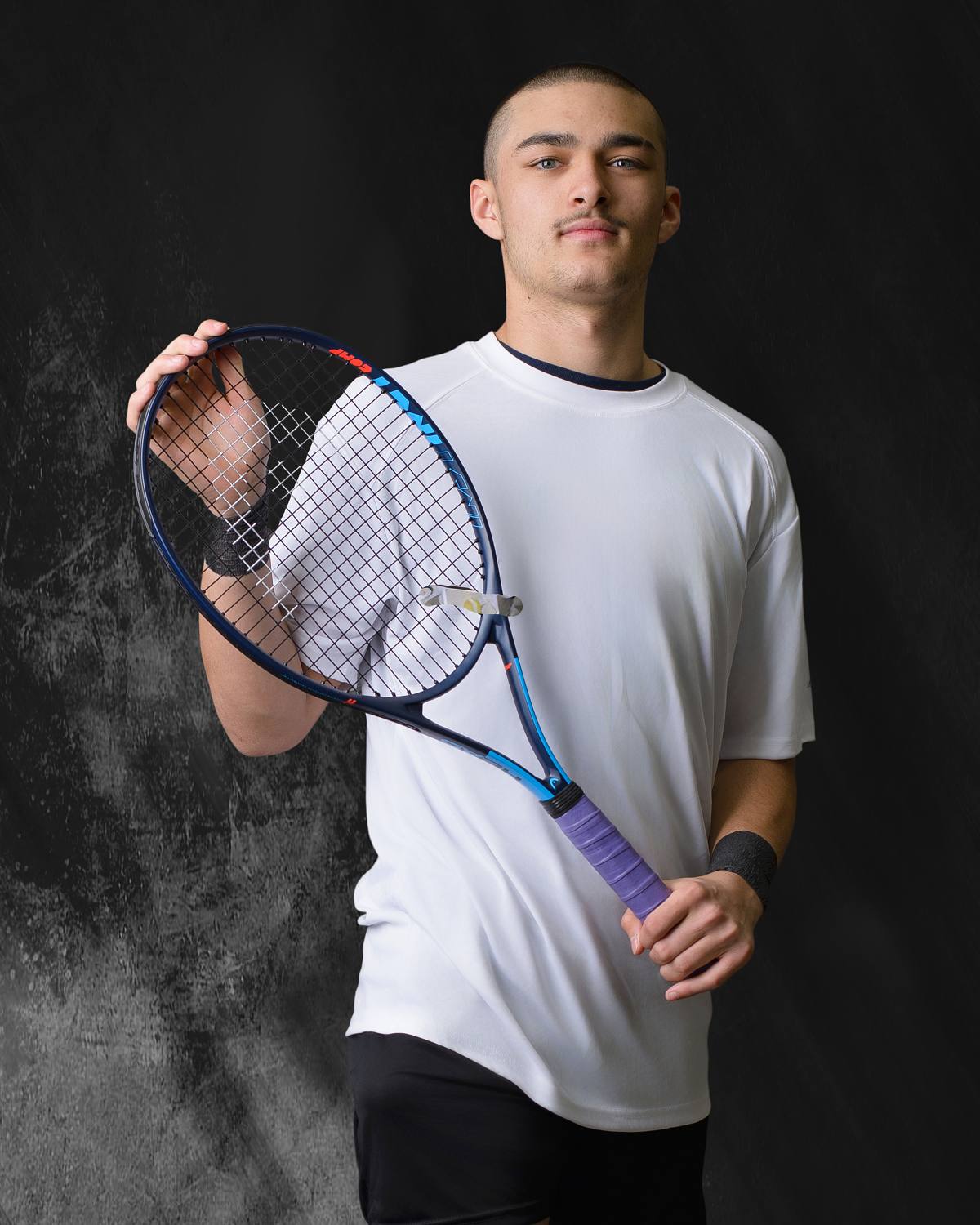A vertical studio portrait by ProClicks Photography of a young male tennis player against a dark, textured background. He has short buzzed hair and a light mustache, wearing a white t-shirt and black shorts. He holds a blue and black tennis racket diagonally across his chest with both hands, looking calmly at the camera.