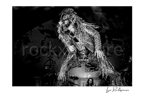 Horizontal black and white image of Rob Zombie performing live on stage with long dreadlocks, a studded fringed jacket, crouched stance, and a handheld microphone Zombie wide horizontal B&W concert photo print
