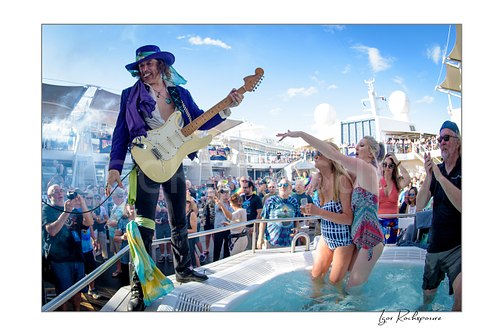 Horizontal color image of Randy Hansen performing with a white guitar on an outdoor ship deck stage beside a small pool with cheering fans around him