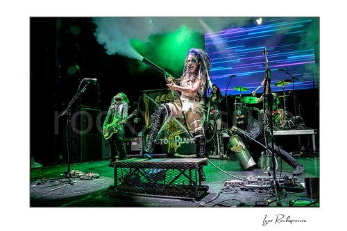 Horizontal color image of Calico Cooper of Beasto Blanco performing live on stage with a smoke cannon, standing on a platform amid green stage smoke while band members appear behind her