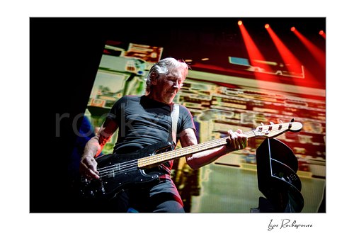Roger Waters looks down while playing a black bass across his waist, with red beams overhead and a patterned stage screen behind him.
