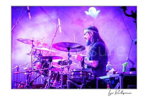 Horizontal color image of Mike Portnoy playing drums in side profile through purple stage smoke during a live performance