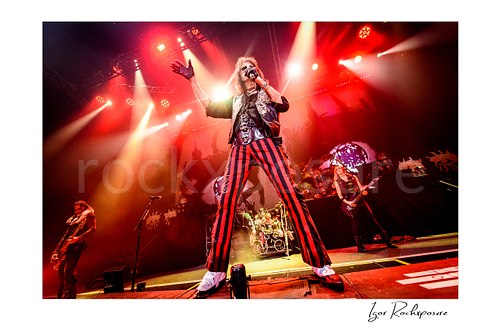 Horizontal color image of Alice Cooper performing live on stage in red striped pants with bright red lighting, raised hand, and the Alice Cooper Band visible behind him