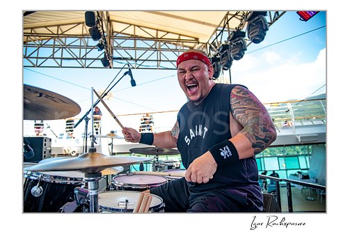 Horizontal color image of Rose Tattoo drummer playing drums onstage in a sleeveless shirt and red bandana during a live performance