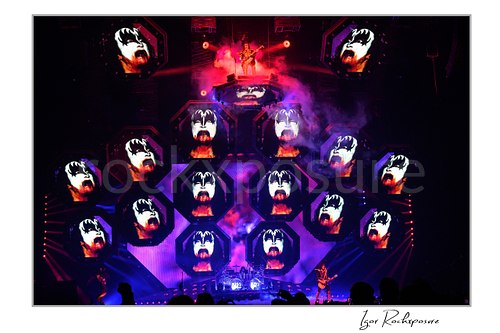 Gene Simmons of KISS standing on an elevated stage platform surrounded by large illuminated Gene Simmons demon face visuals across the stage screens under red and purple concert lighting