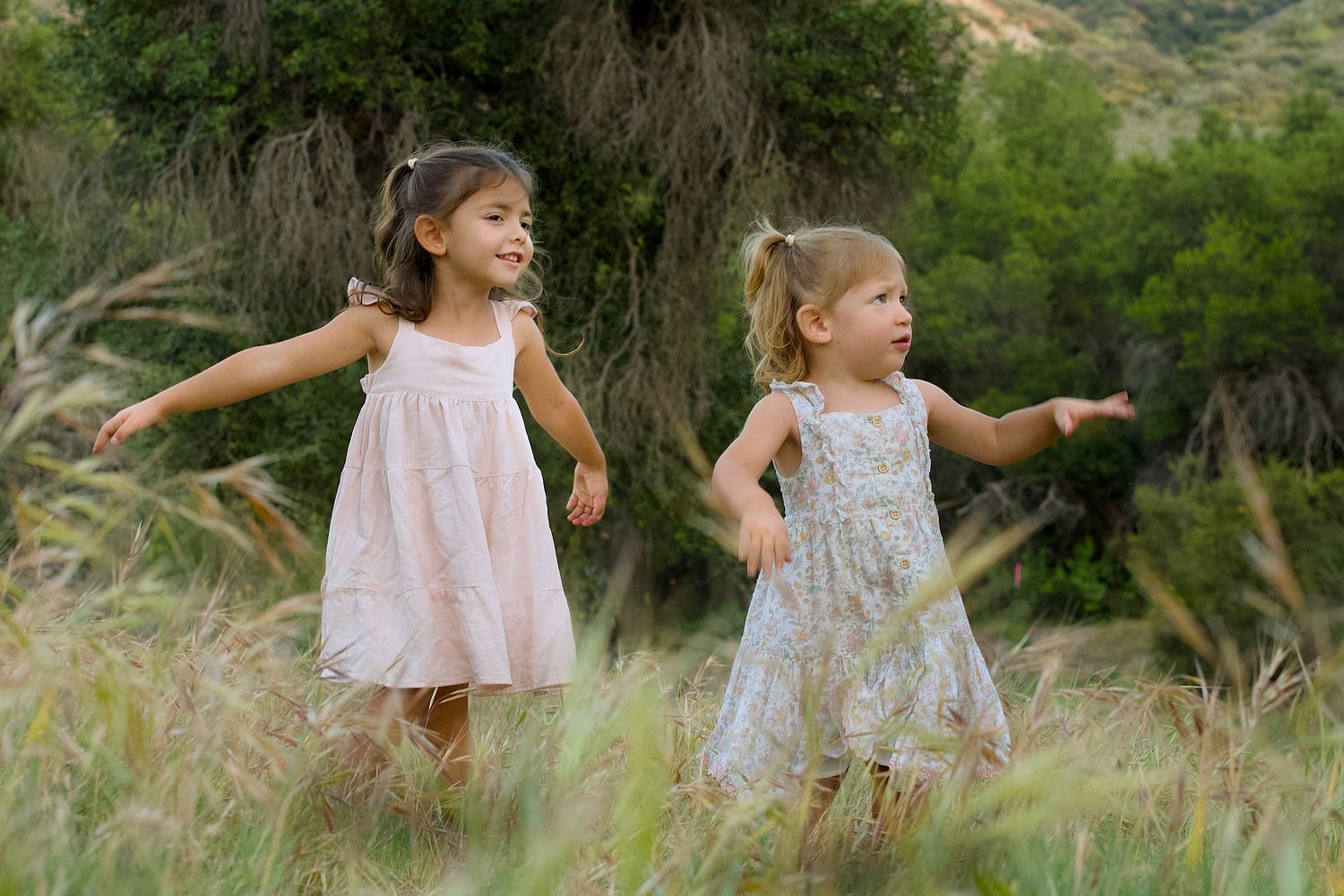 Two sisters in light dresses running through tall grass.