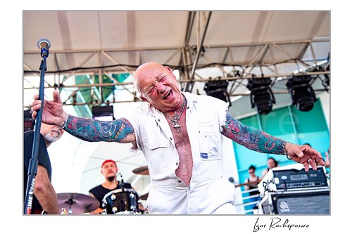 Horizontal color image of Angry Anderson of Rose Tattoo performing onstage in a white sleeveless outfit with arms outstretched and the drummer visible behind him