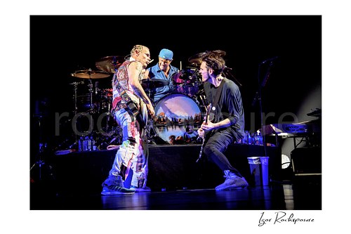 Flea and Josh Klinghoffer face each other with bass and guitar before Chad Smith’s drum kit, blue light reflecting across the stage floor.